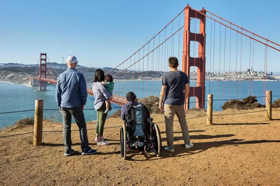 Eine Gruppe von Menschen, darunter eine Person im Rollstuhl, ist von hinten zu sehen, wie sie von den Marin Headlands aus auf die Golden Gate Bridge blicken.